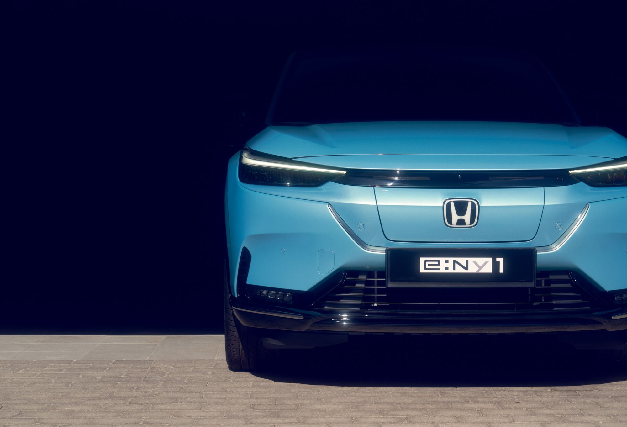 Front view of a light blue Honda e:NY1 electric vehicle, showing the sleek headlights, Honda logo, and illuminated license plate against a dark background.